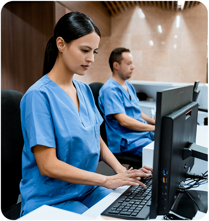 A healthcare professional wearing blue scrubs is seated at a desk, typing on a computer in a modern medical facility. Another professional in blue scrubs is seated in the background, also working at a computer. The setting is well-lit with a clean and professional appearance. A healthcare professional wearing blue scrubs is seated at a desk, typing on a computer in a modern medical facility. Another professional in blue scrubs is seated in the background, also working at a computer. The setting is well-lit with a clean and professional appearance.